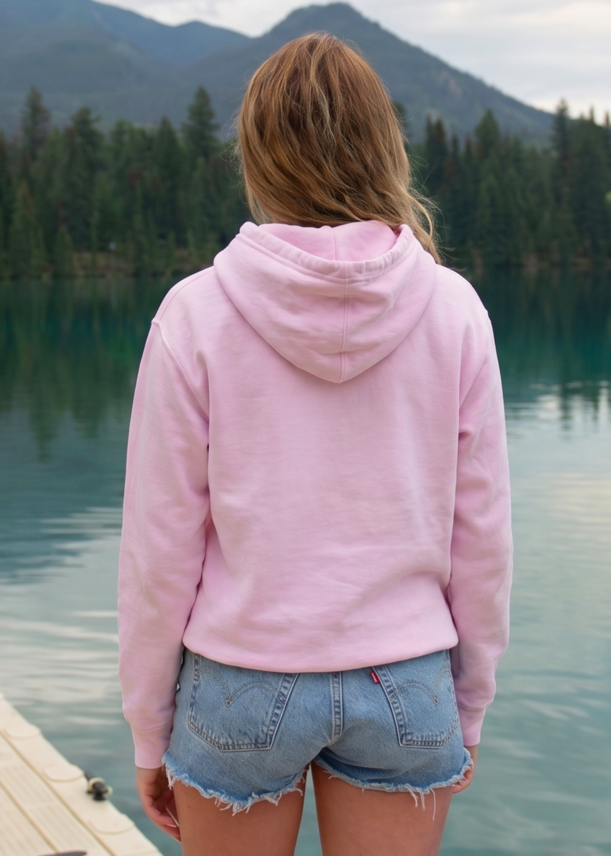 Person wearing a pink hoodie and denim shorts standing by a lake with mountains in the background