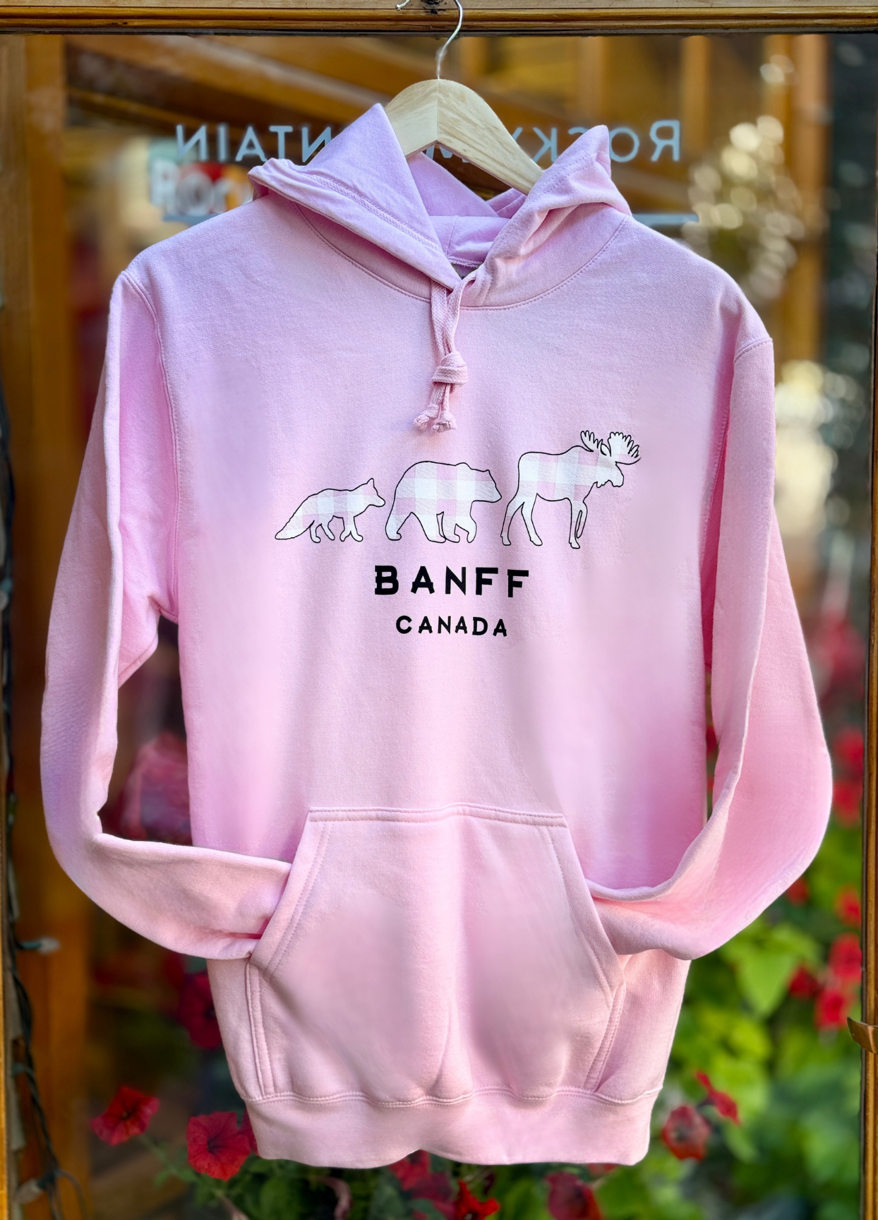 Pink hoodie with 'Banff Canada' text and moose graphics on a hanger against a blurred floral background.