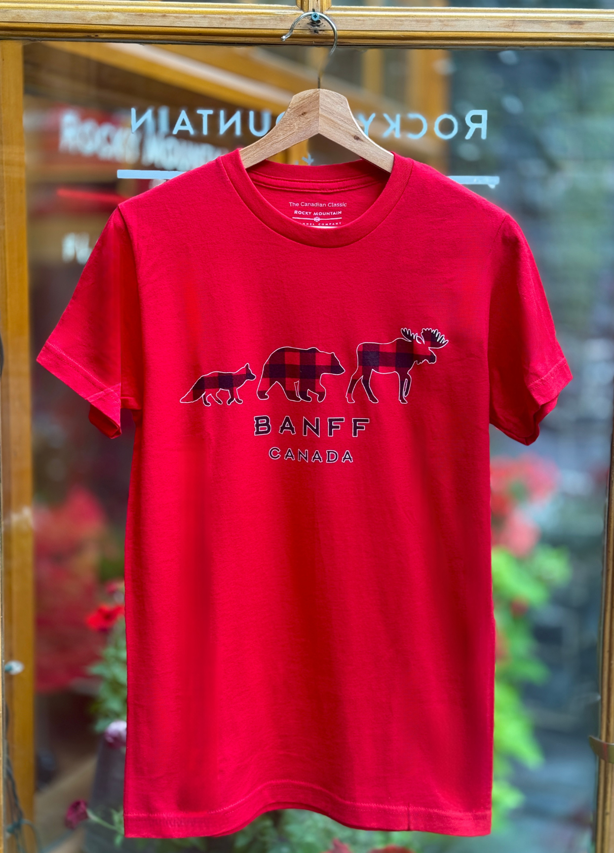 Red t-shirt with 'Banff Canada' design on a hanger in front of a store window.