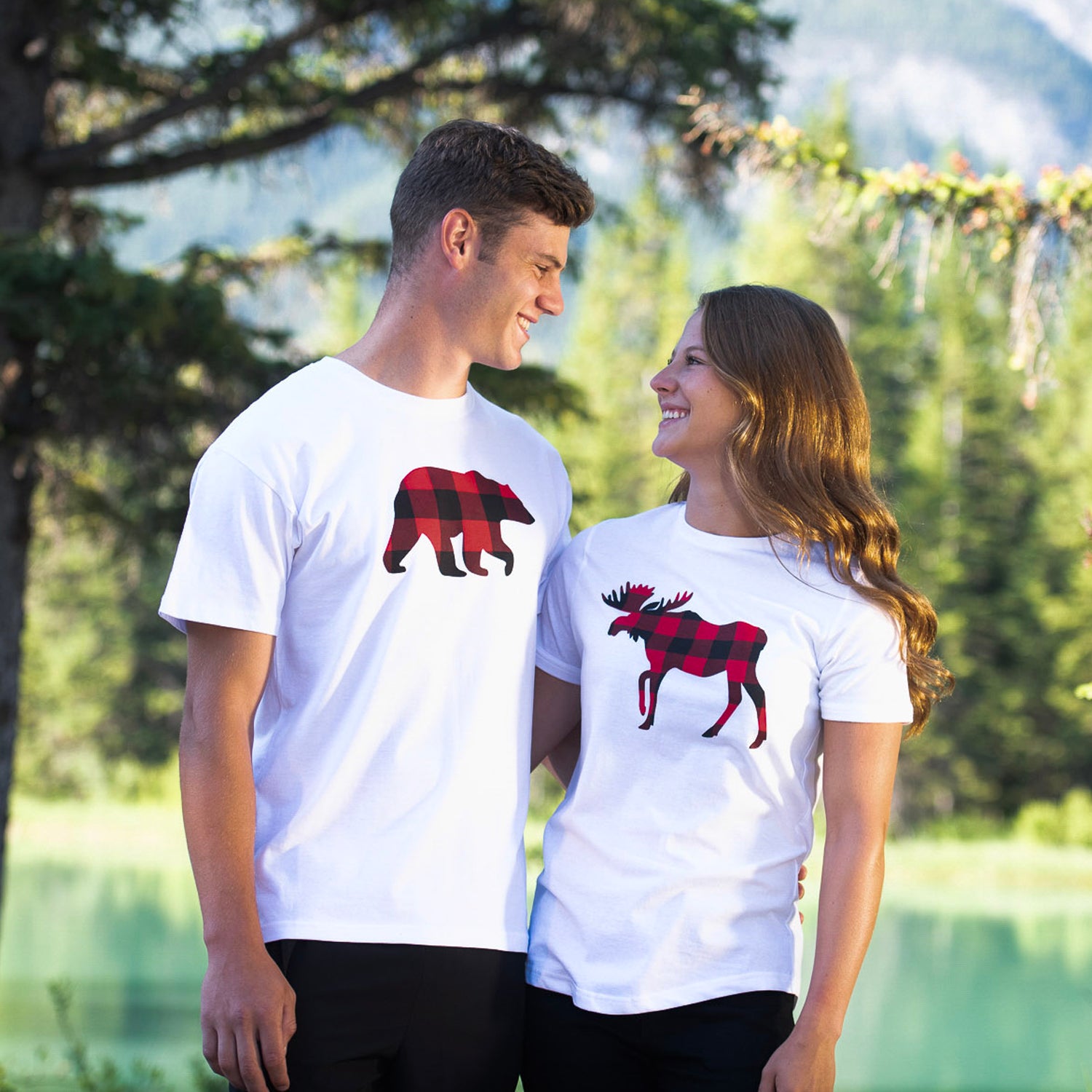 Two people wearing white t-shirts with red and black buffalo check animal designs in a natural setting.