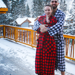 Two people in matching red and black buffalo check robes standing on a snowy deck with a mountainous background.