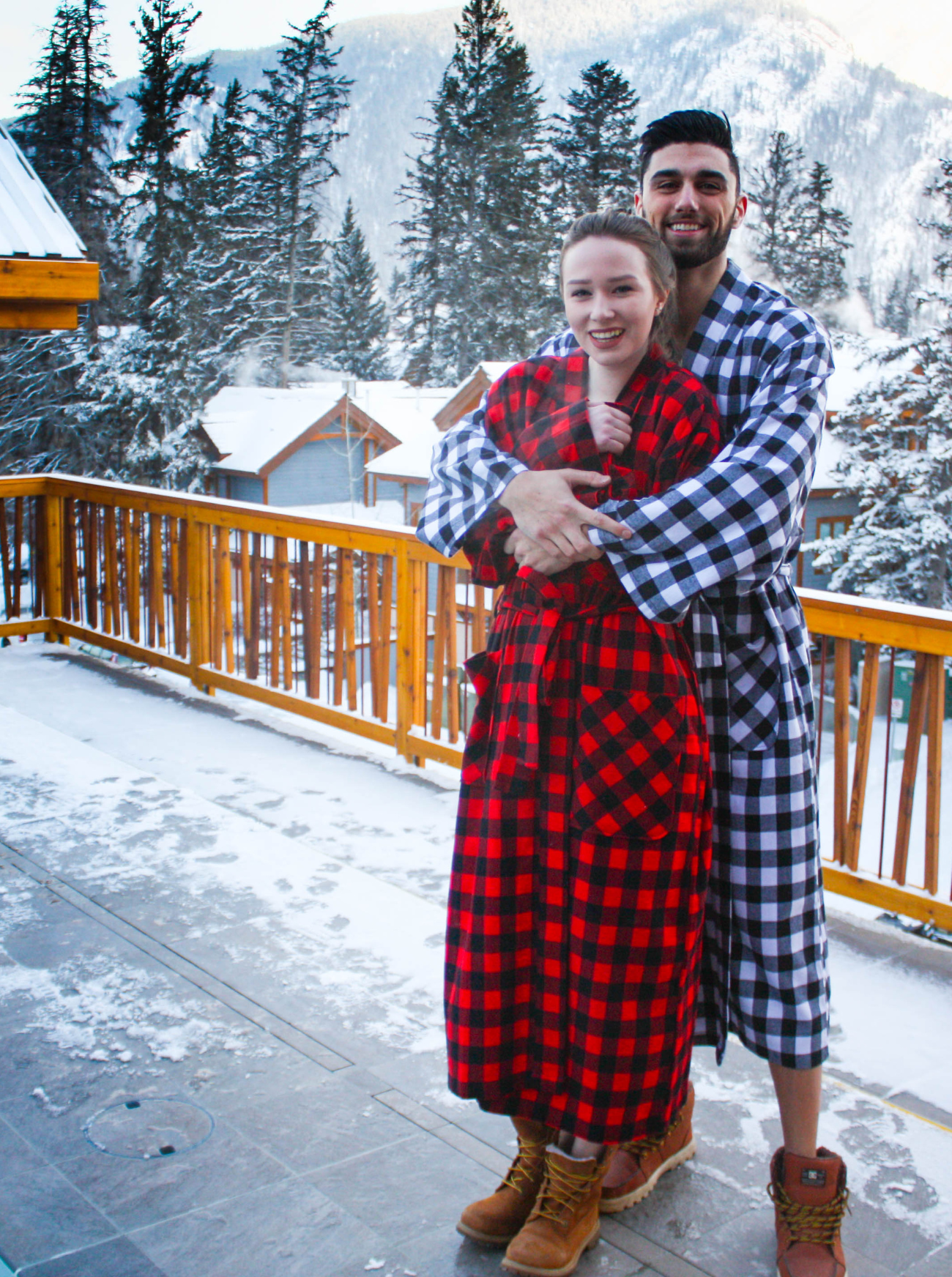Two people in matching red and black buffalo check robes standing on a snowy deck with a mountainous background.