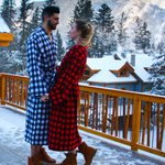 Two people in buffalo check robes standing on a snowy deck with mountains in the background