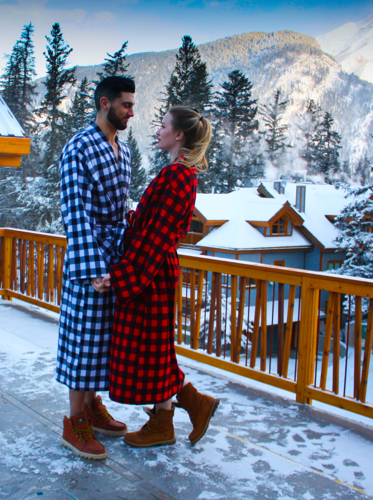 Two people in buffalo check robes standing on a snowy deck with mountains in the background