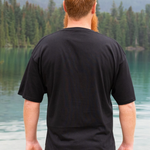 Man wearing a black t-shirt standing by a lake with mountains in the background