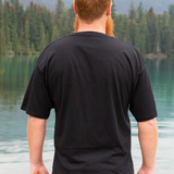 Man wearing a black t-shirt standing by a lake with mountains in the background