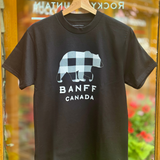 Black t-shirt with a bear graphic and 'Banff Canada' text on a hanger against a blurred indoor background.
