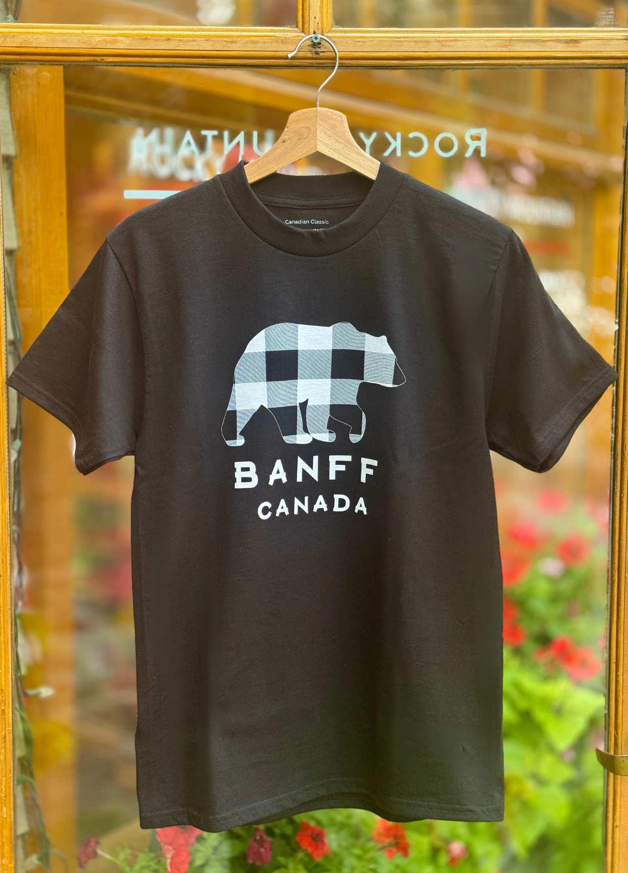 Black t-shirt with a bear graphic and 'Banff Canada' text on a hanger against a blurred indoor background.