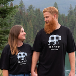 Two people wearing black Banff t-shirts standing in a forested area with mountains in the background.