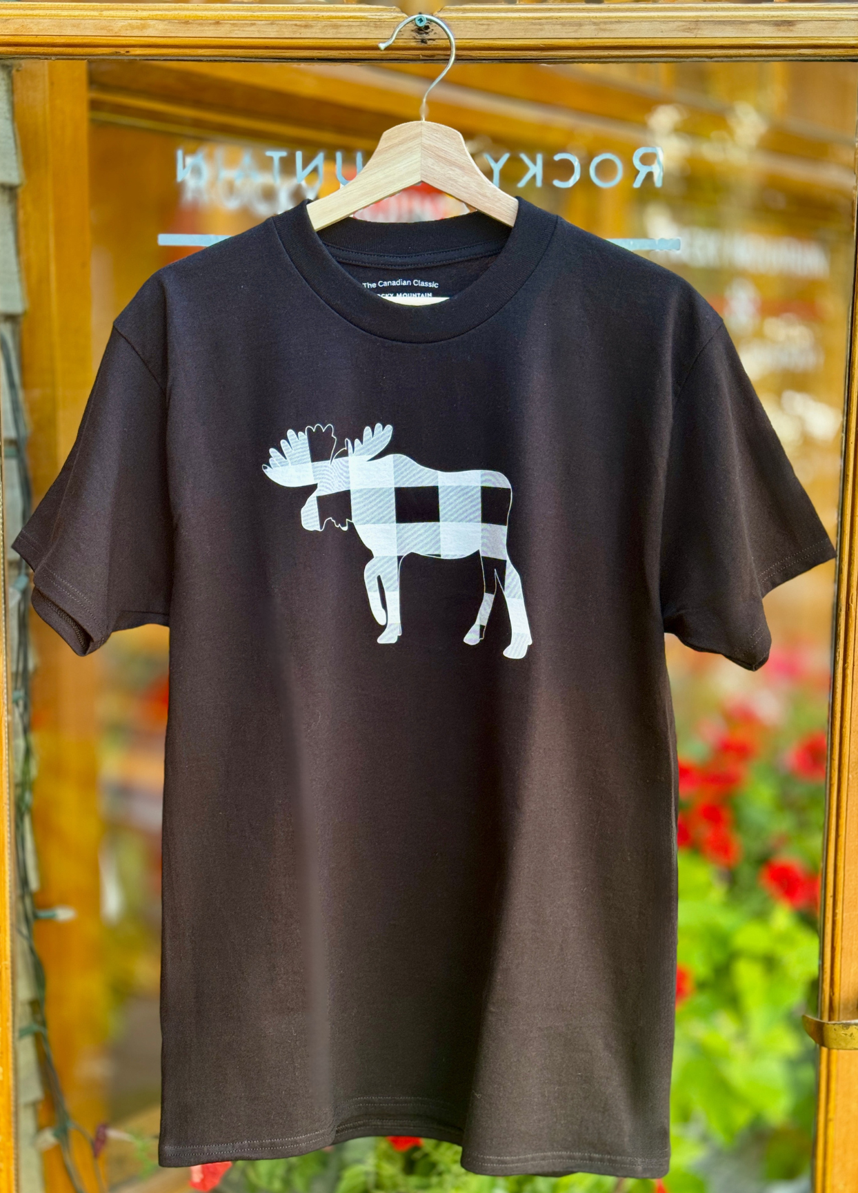 Black t-shirt with a white moose graphic on a hanger in front of a store window.