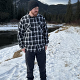 A man wearing a black and white plaid padded flannel jacket, standing near a snowy landscape.