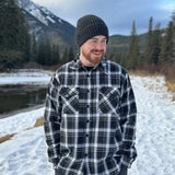 A man wearing a black and white plaid padded flannel jacket, standing near a snowy landscape.