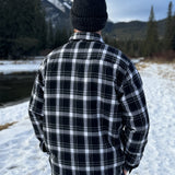 Back view of a man wearing a black and white plaid padded flannel jacket, standing near a snowy landscape.