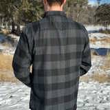 Back view of man wearing a long-sleeved black and grey buffalo check flannel shirt with two breast pockets and a button cuff.