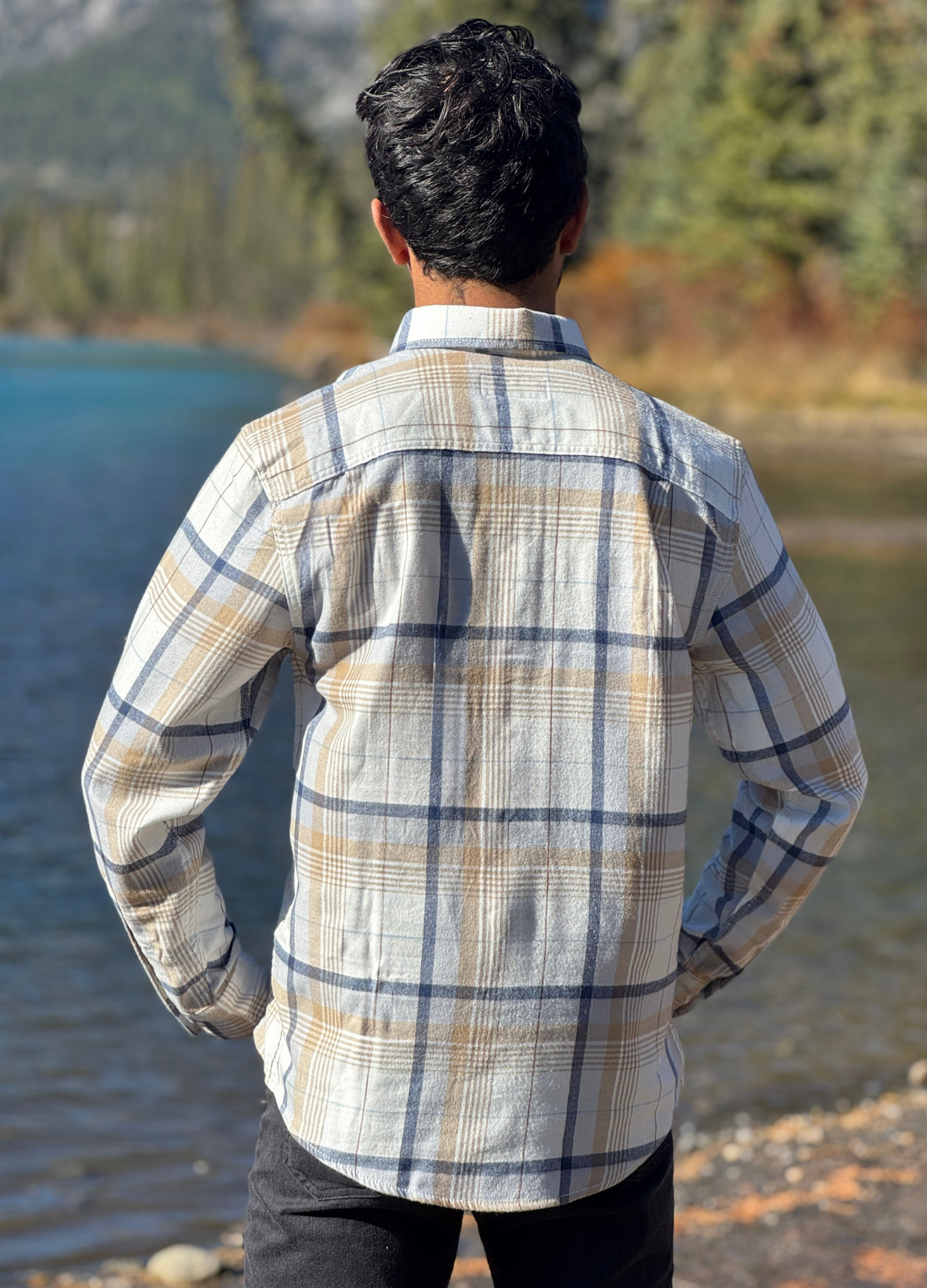 Person wearing a plaid flannel shirt by a river with mountains in the background in Banff National Park