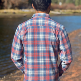 Man wearing a plaid flannel shirt standing by a body of water with trees in the background