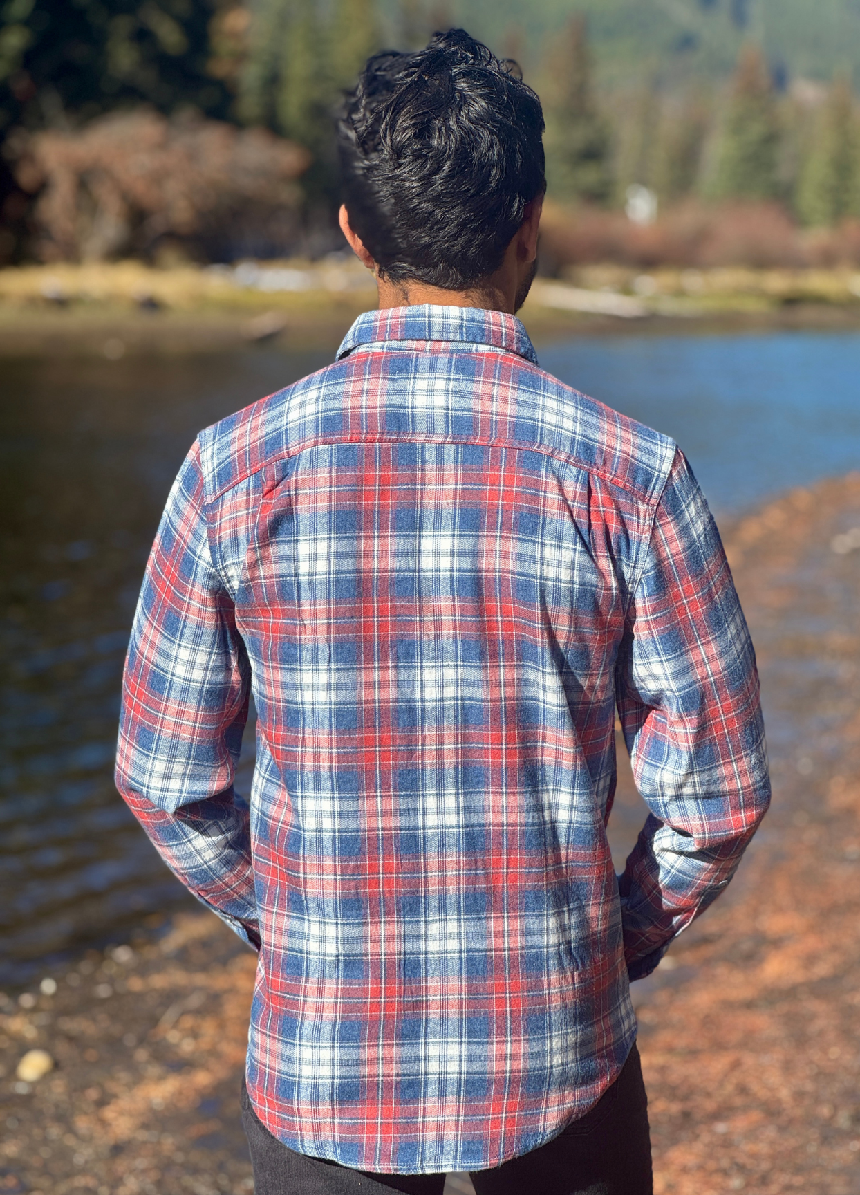 Man wearing a plaid flannel shirt standing by a body of water with trees in the background
