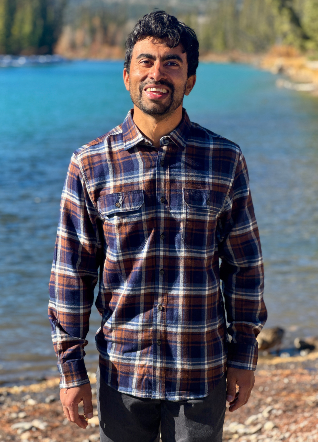 Man wearing a plaid flannel shirt standing by a river with trees in the background in Banff National Park