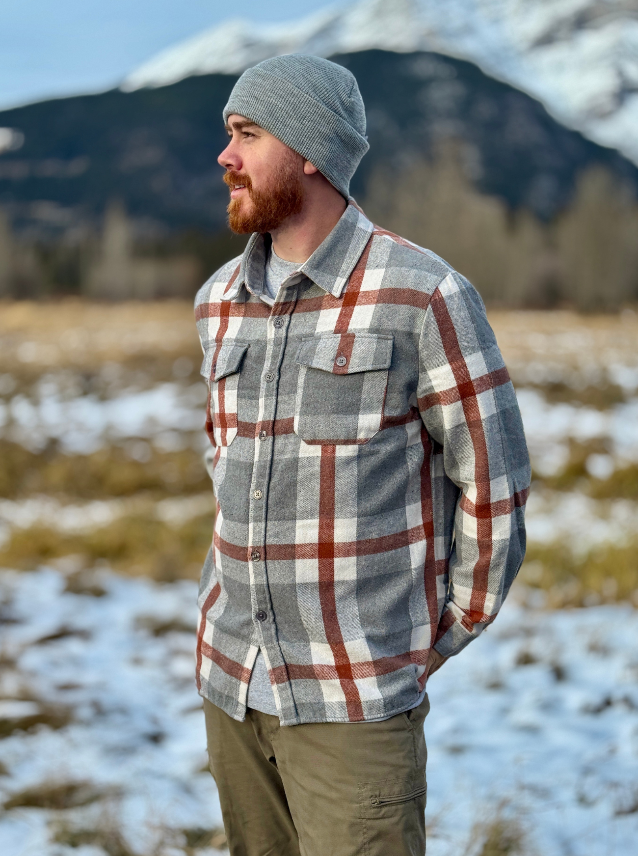 Man wearing a plaid shirt and beanie standing in a snowy landscape with mountains in the background