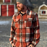 Man wearing a plaid flannel shirt and knit cap standing outdoors with a barn in the background