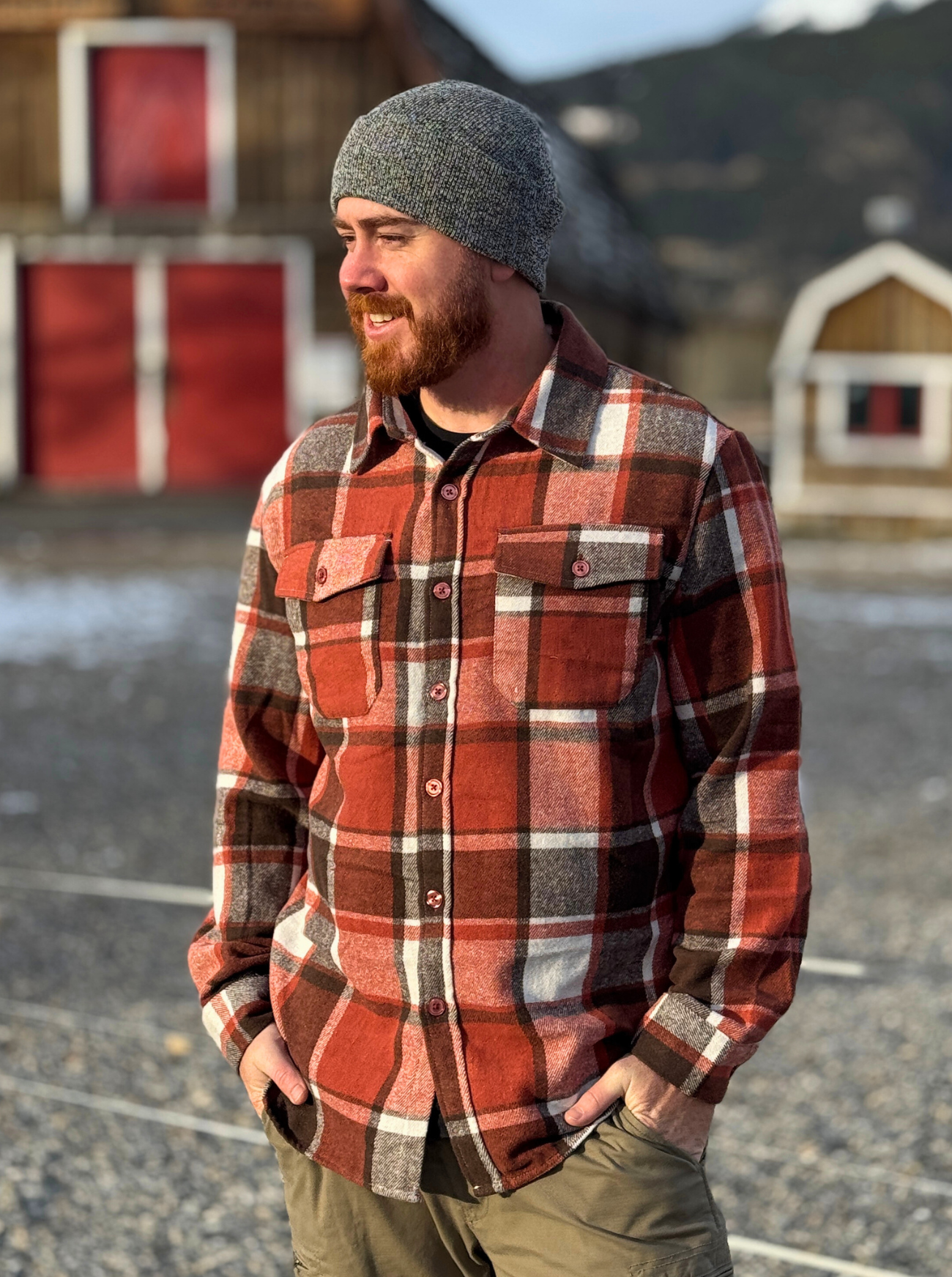 Man wearing a plaid flannel shirt and knit cap standing outdoors with a barn in the background