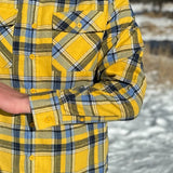 A man wearing a yellow and blue large plaid long-sleeved shirt standing outdoors.
