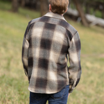 Back view of a person wearing a brown wool blend flannel jacket with herringbone weave, paired with blue jeans, standing in a natural outdoor setting.