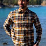 Man wearing a plaid flannel shirt standing by a body of water with trees in the background in Banff National Park