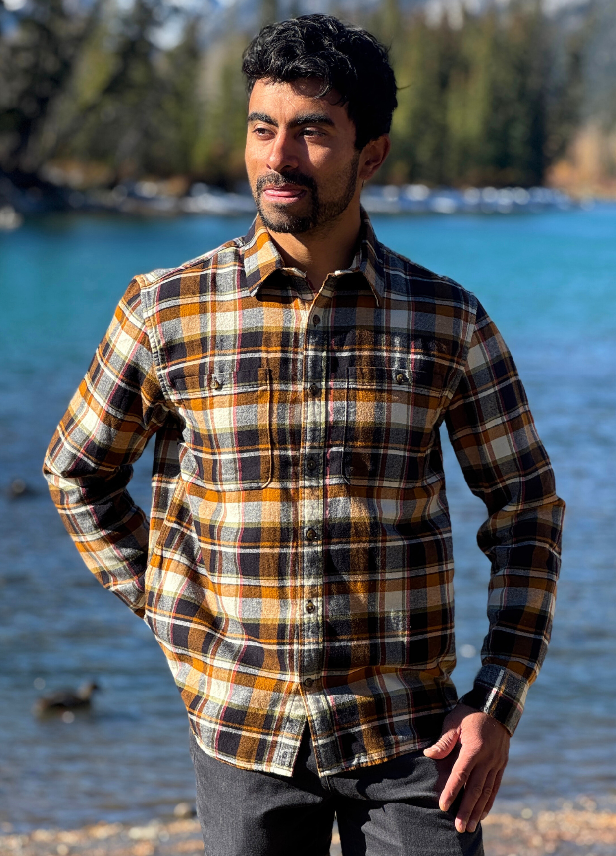 Man wearing a plaid flannel shirt standing by a body of water with trees in the background in Banff National Park