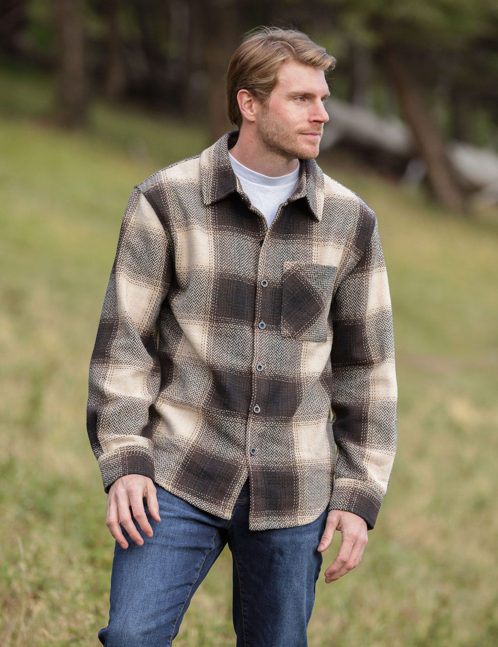 A person wearing a brown wool blend flannel jacket with herringbone weave, paired with blue jeans, standing in a natural outdoor setting.