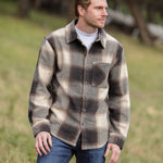 A person wearing a brown wool blend flannel jacket with herringbone weave, paired with blue jeans, standing in a natural outdoor setting.