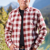 An older man with short grey hair wearing a burgundy and white buffalo check flannel shirt and blue jeans, standing near a body of water.