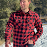 A man wearing a red and black buffalo check flannel shirt with two flap breast pockets and a button cuff, standing outdoors. This is an action shot, he is walking.
