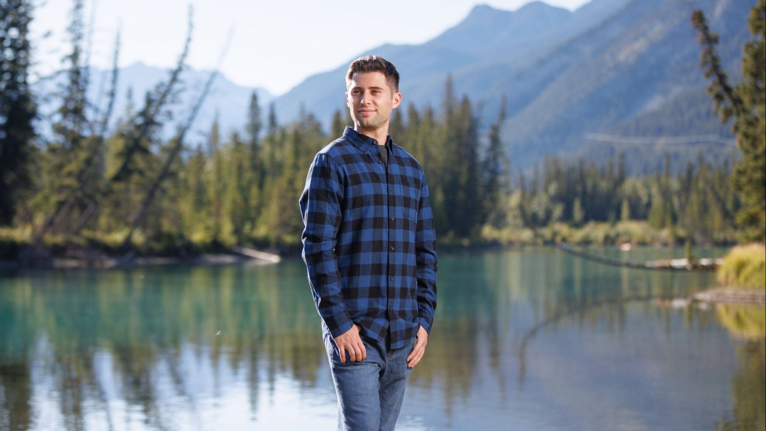 Man standing in front of a river in Banff National Park. Wearing our Canadian Classic Blue Buffalo Check Mens Shirt. His hands are in his front pockets and he has styled the shirt with blue jeans.