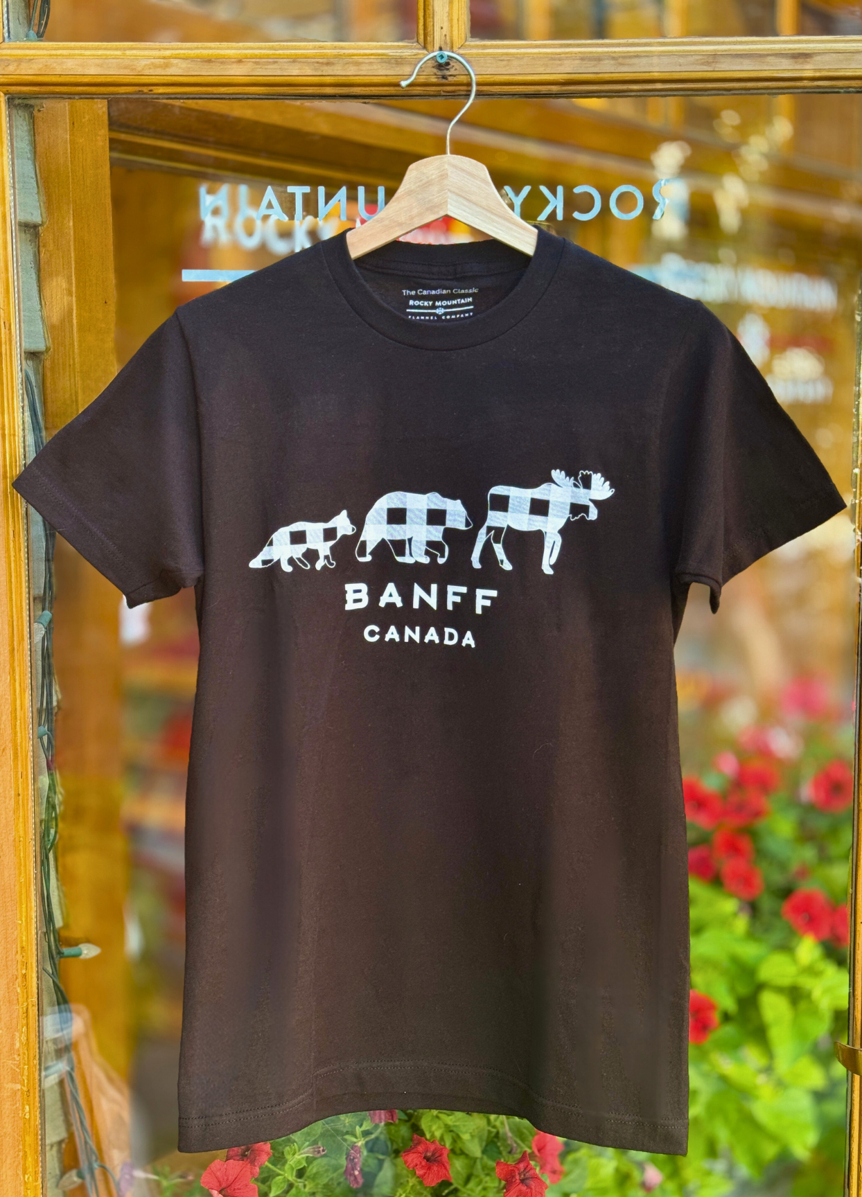 Black t-shirt with white graphic design and text 'Banff Canada' on a hanger against a blurred indoor background.