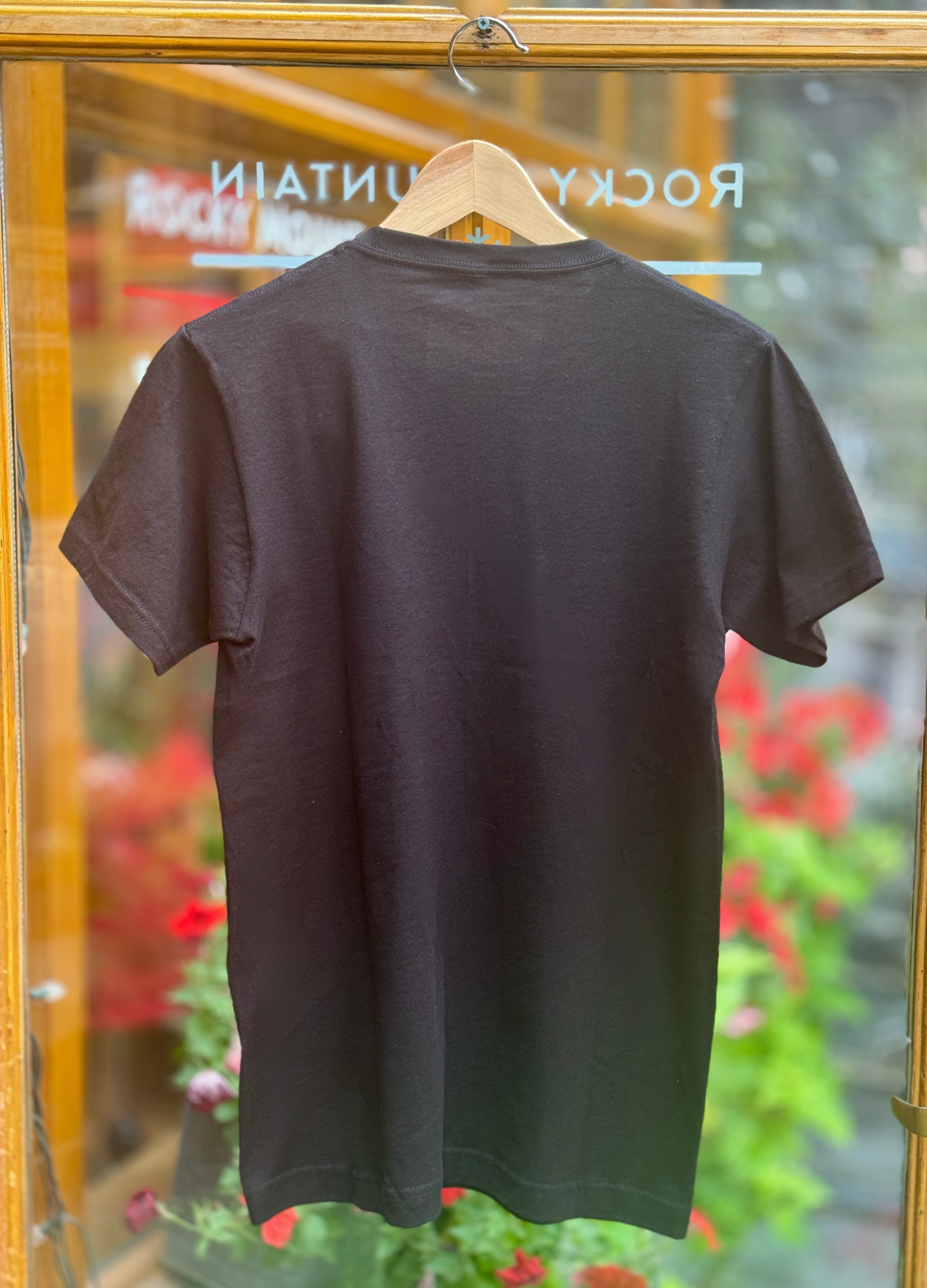 Black t-shirt on a hanger with a blurred background of flowers and a store entrance.