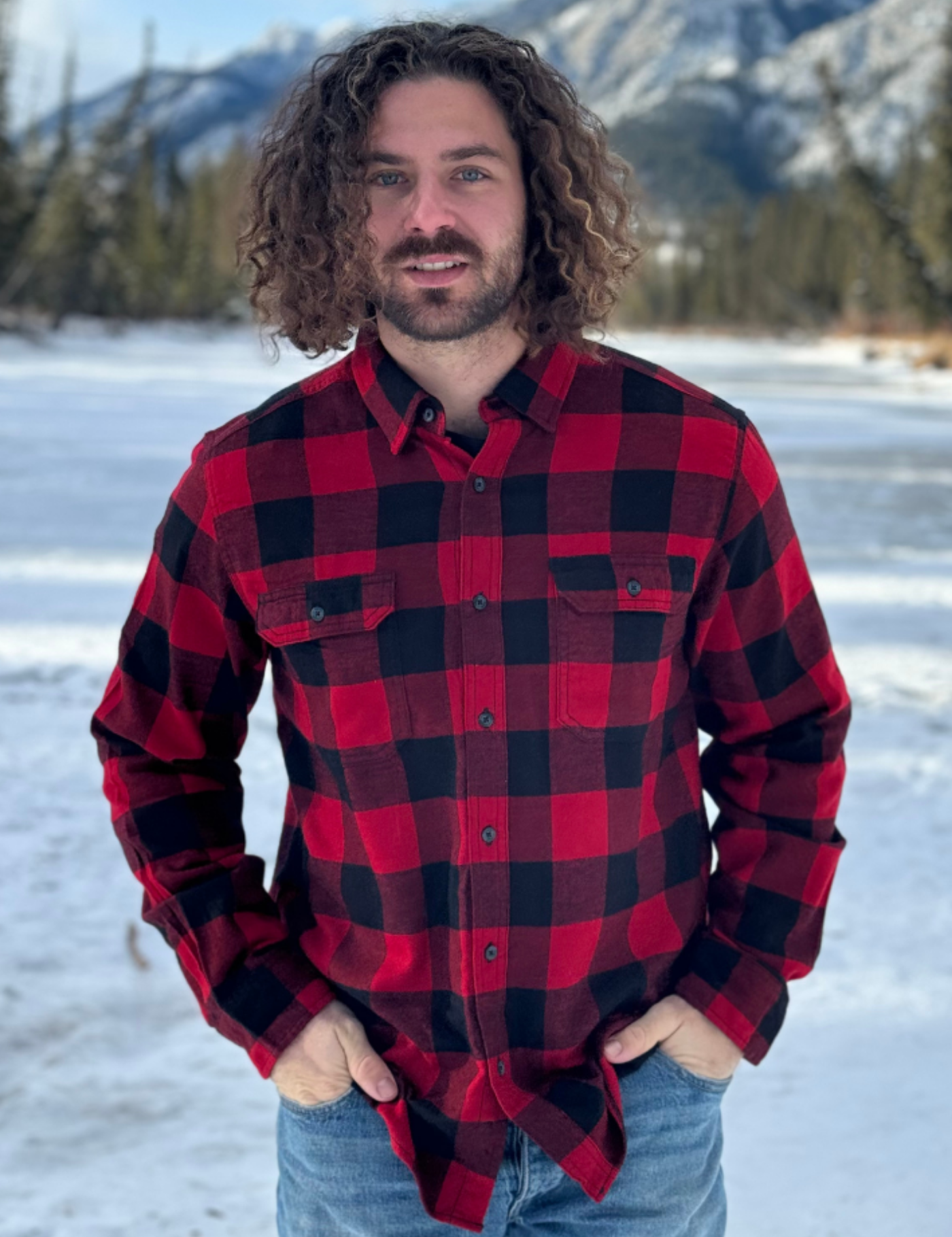 Man wearing a red and black buffalo check shirt standing in a snowy landscape with mountains in the background in Banff, Canada