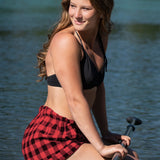 A woman sitting by a lake wearing red and black checkered short shorts with a twill drawstring and side pockets.