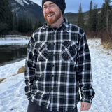 A man wearing a black and white plaid padded flannel jacket, standing near a snowy landscape.