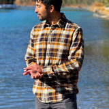 Man wearing a plaid flannel shirt standing by a lake with mountains in the background