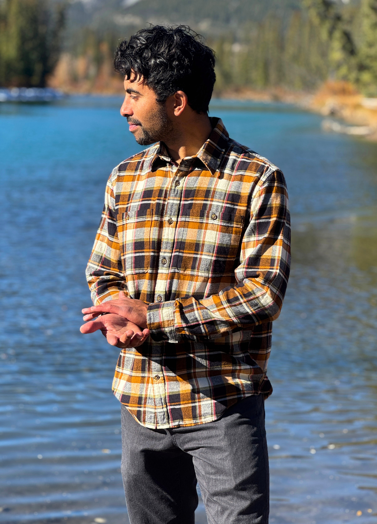 Man wearing a plaid flannel shirt standing by a lake with mountains in the background