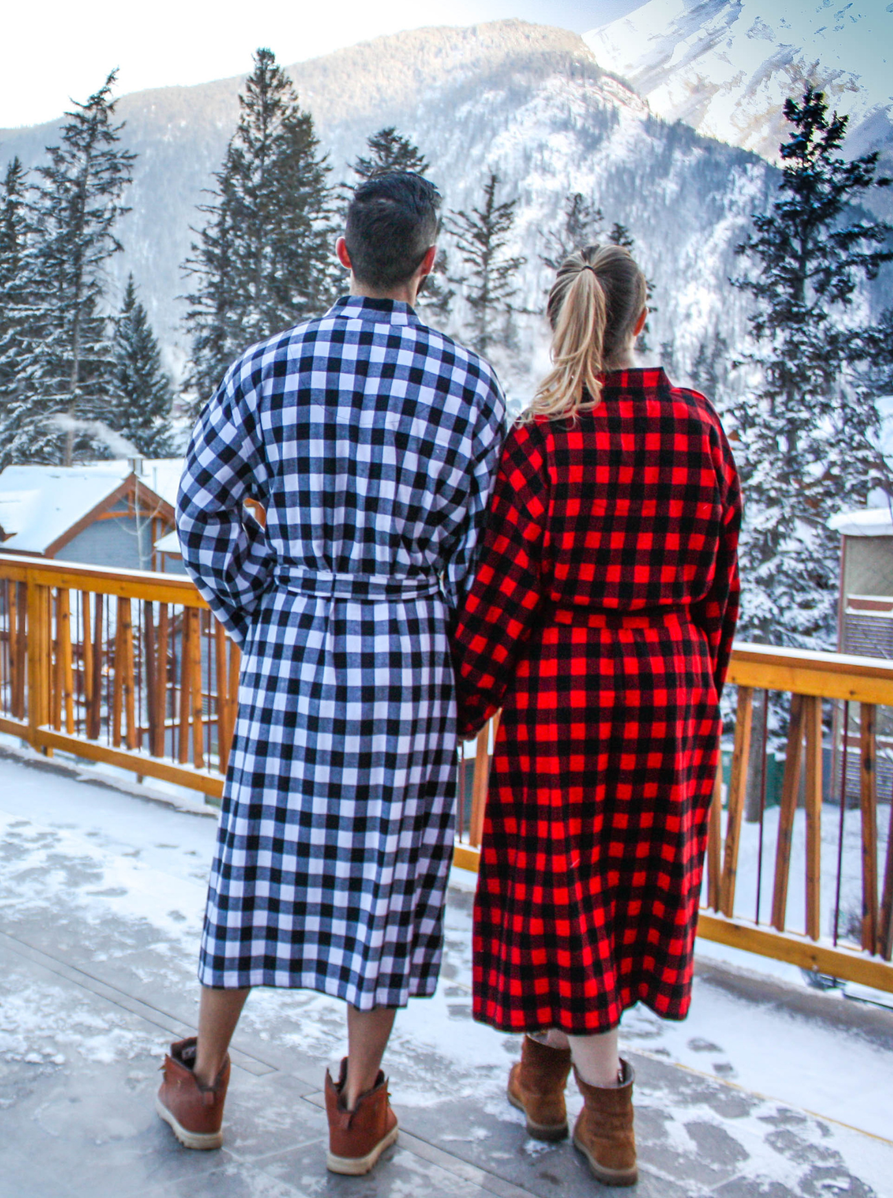 Two people wearing buffalo check robes standing on a snowy deck with mountains in the background in Banff, Canada