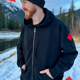 A person wearing a full zip black hoody with a kangaroo pocket, standing outdoors with snow and trees in the background.