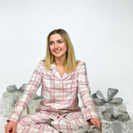 Woman wearing pink plaid pajamas sitting on a white surface with wrapped gifts in the background.