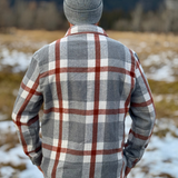 Person wearing a plaid flannel shirt and gray beanie standing in a snowy field with mountains in the background