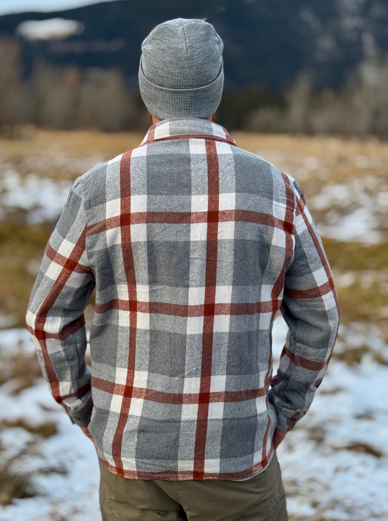 Person wearing a plaid flannel shirt and gray beanie standing in a snowy field with mountains in the background