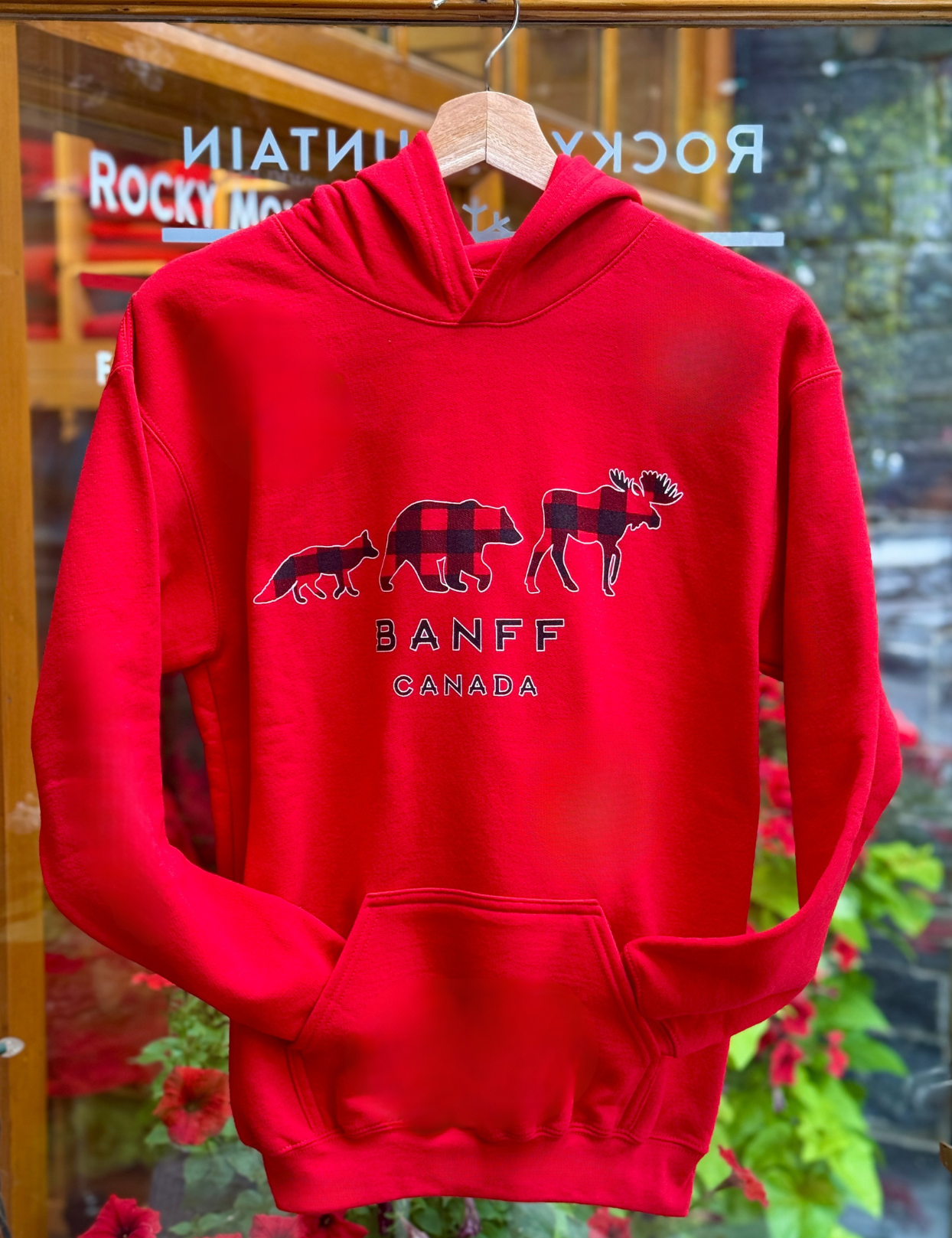 Kids red hoodie with moose, bear and fox graphic and 'Banff Canada' text on a hanger in front of a store window.