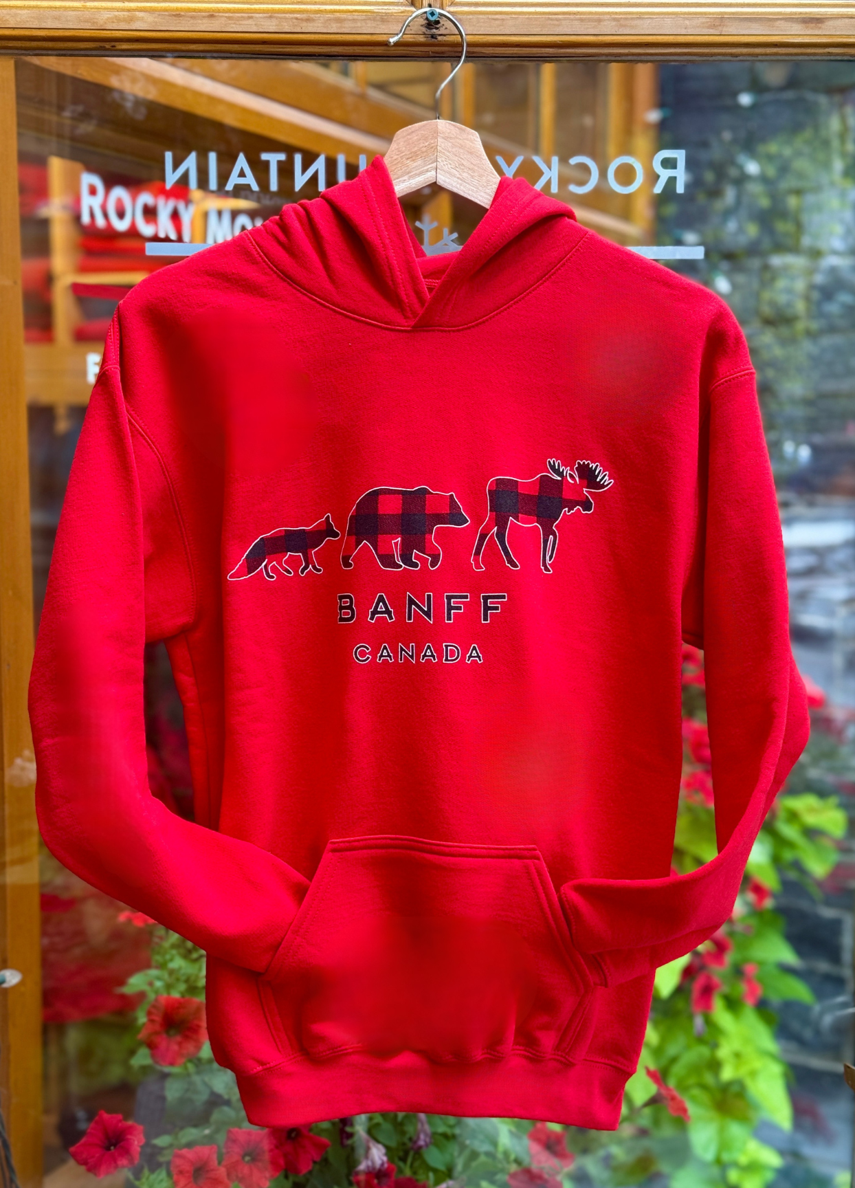 Kids red hoodie with moose, bear and fox graphic and 'Banff Canada' text on a hanger in front of a store window.