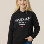 A young girl with blonde hair smiling while wearing a black pullover hoodie with a white Banff Canada graphic and blue jeans, set against a solid light beige background.