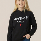A young girl with blonde hair smiling while wearing a black pullover hoodie with a white Banff Canada graphic and blue jeans, set against a solid light beige background.